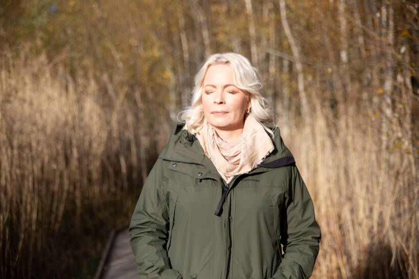 Kuvassa haastateltava Jenni Lehtimäki poseeraa kaislikossa kasvot auringon suuntaan, silmät kiinni nauttien. 
